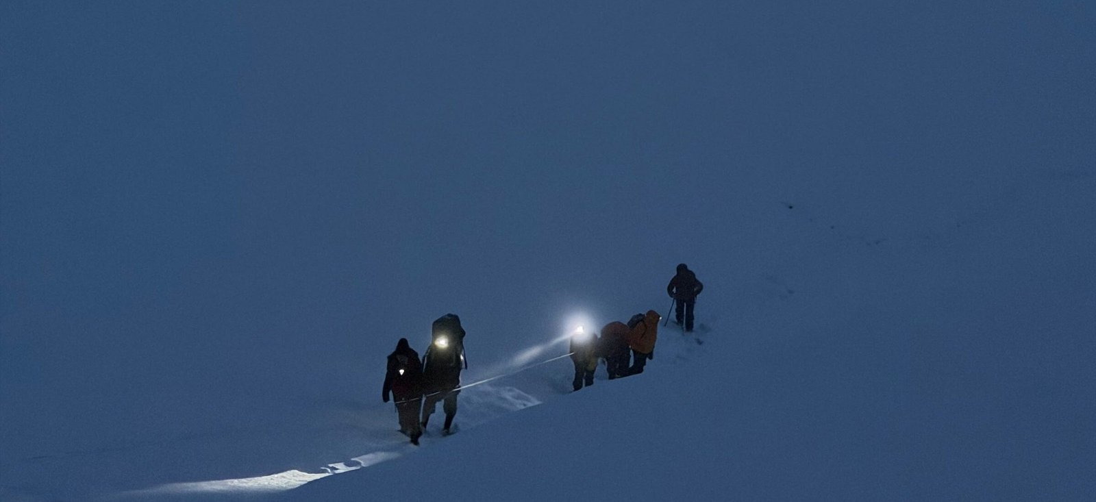 Climbers moving roped across snow before dawn, one headlamp lit, on the way to the pass